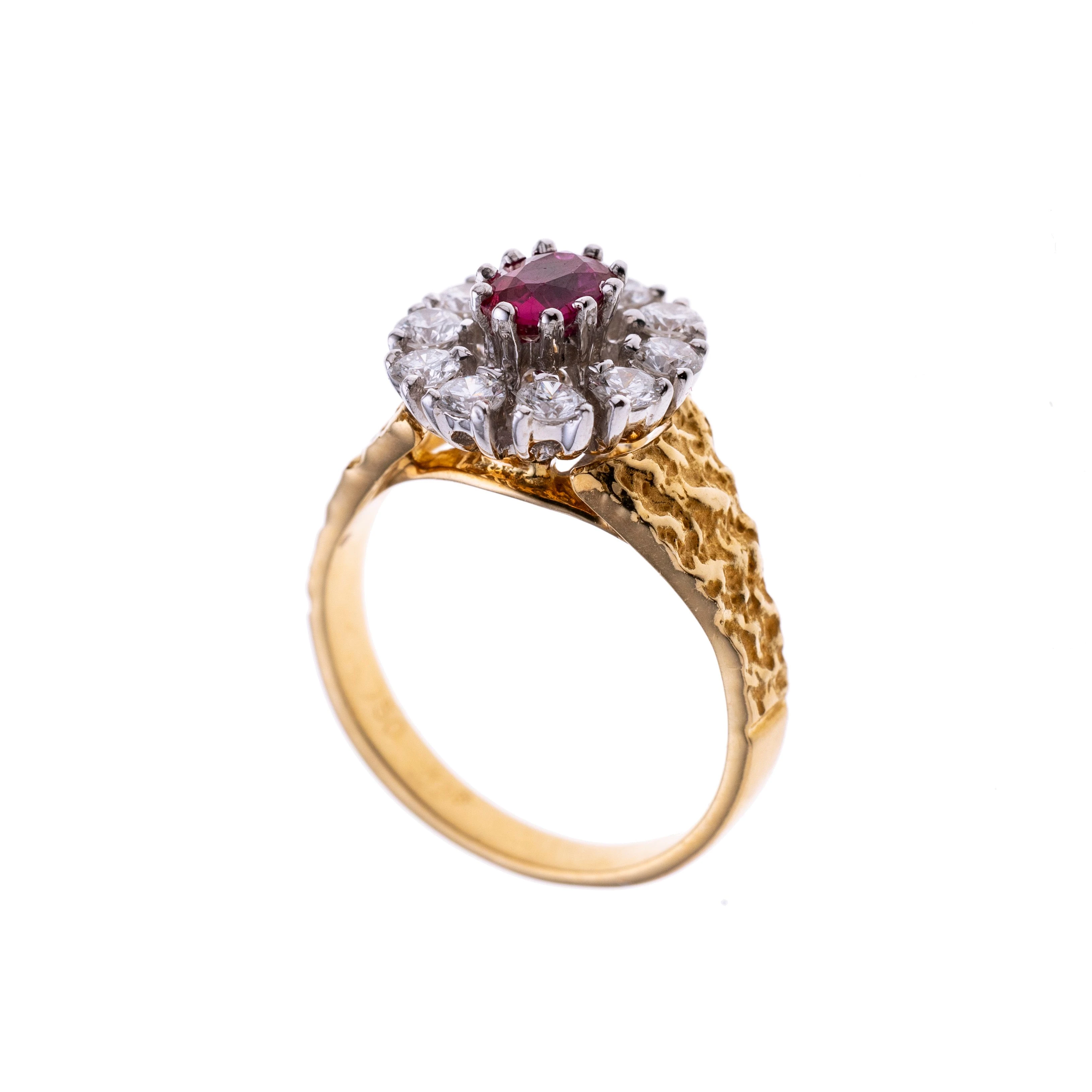 Ein ovaler Rubin karmoisiert von 10 Brillanten. Auf einer gelbgoldenen Ringschiene mit strukturierten Ringschultern.