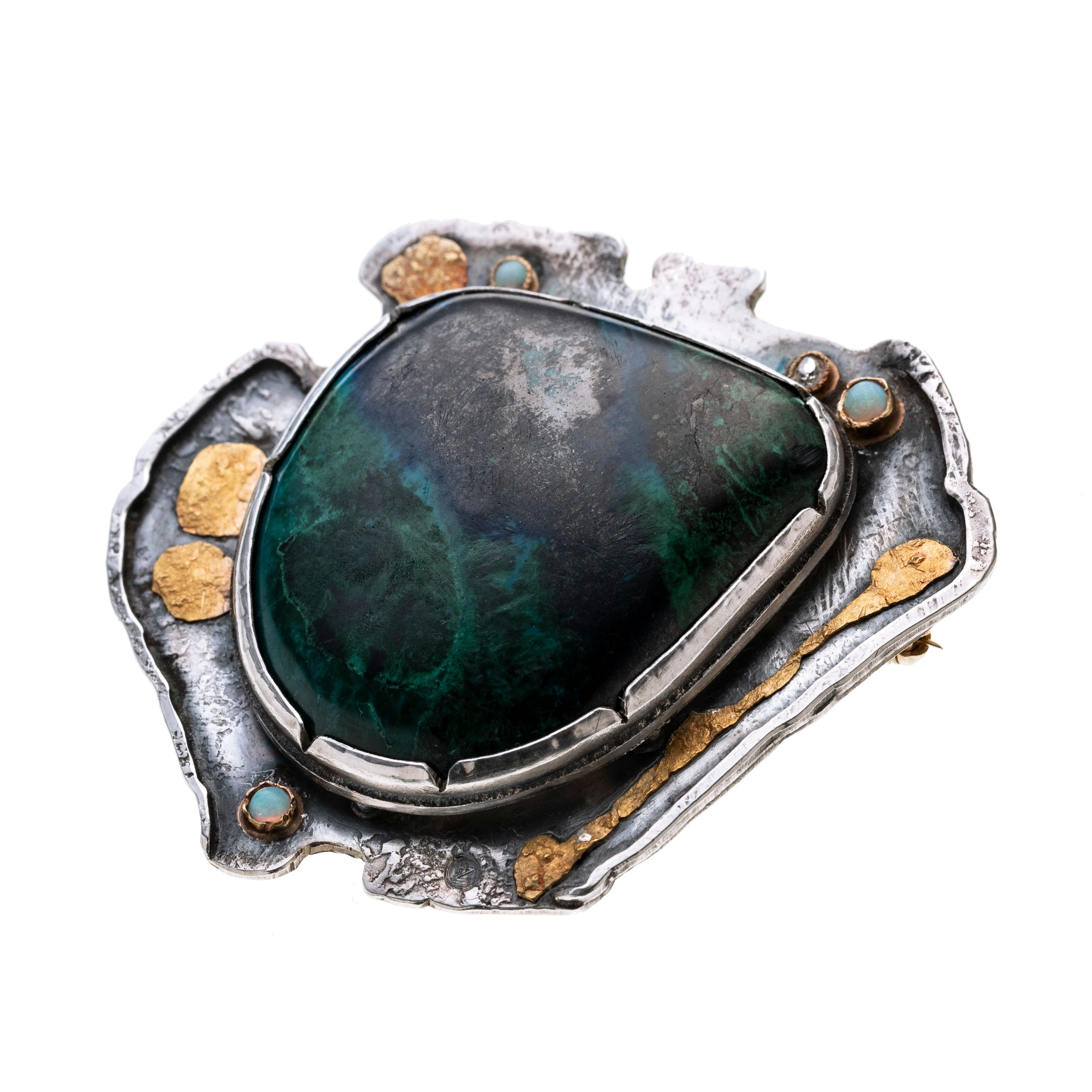 Ein Chrysokoll-Cabochon in in einer durchbrochenen Zargenfassung, mittig auf einer amorph gestalteten Form.  Vier weitere goldene Zargenfassungen sind auf der Fläche aufgebracht, in denen drei Opale und ein Diamant gefasst worden sind. Goldene „Spritzer“ sind zusätzlich aufgelötet.