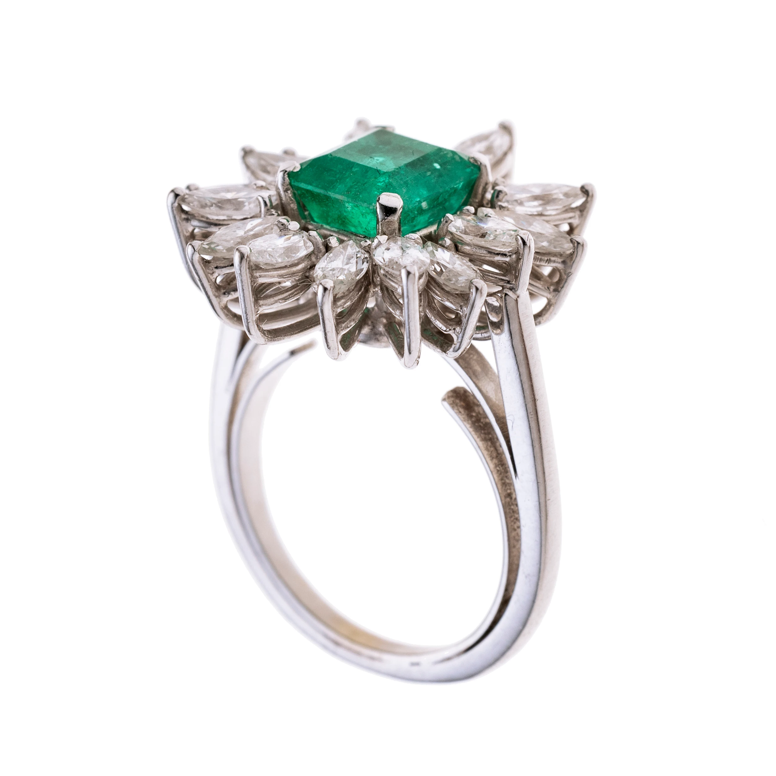 Ein quadratischer Smaragd im Emeraldcut umgeben von 16 Diamantnavetten an einer zarten Ringschiene mit Spange.